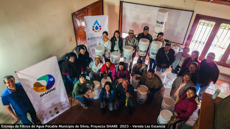 A view down from a stairwell at a group of community members stood together and smiling below, holding their new water filters